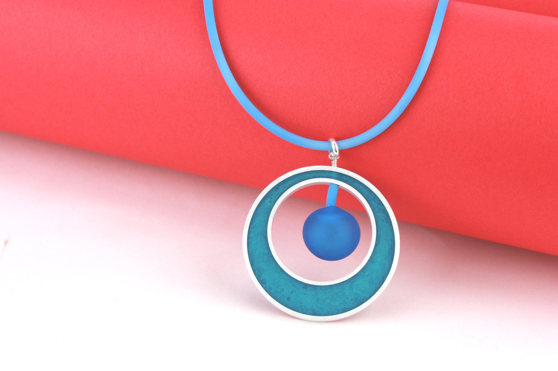 The Glory Necklace in Aqua, featuring a handcrafted pendant made from eco-resin, salvaged aluminum, and frosted blown glass, suspended on a fully adjustable silicone cord.