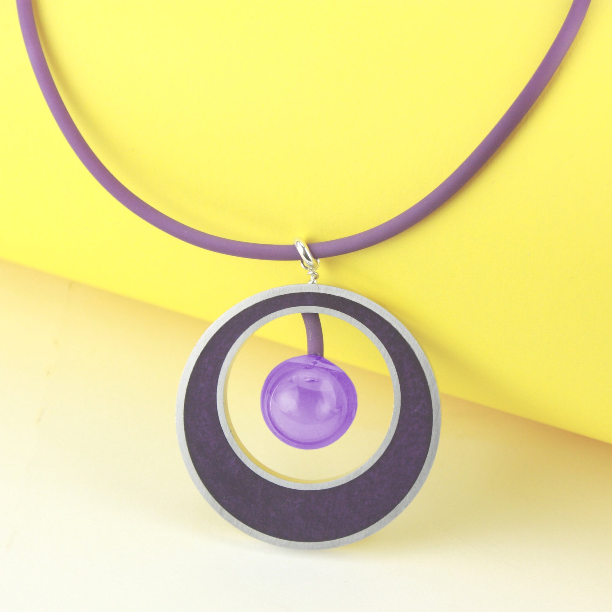 The Glory Necklace in Color-Changing Purple, featuring a handcrafted pendant made from eco-resin, salvaged aluminum, and a blown glass bead that shifts from purple to blue under fluorescent light.