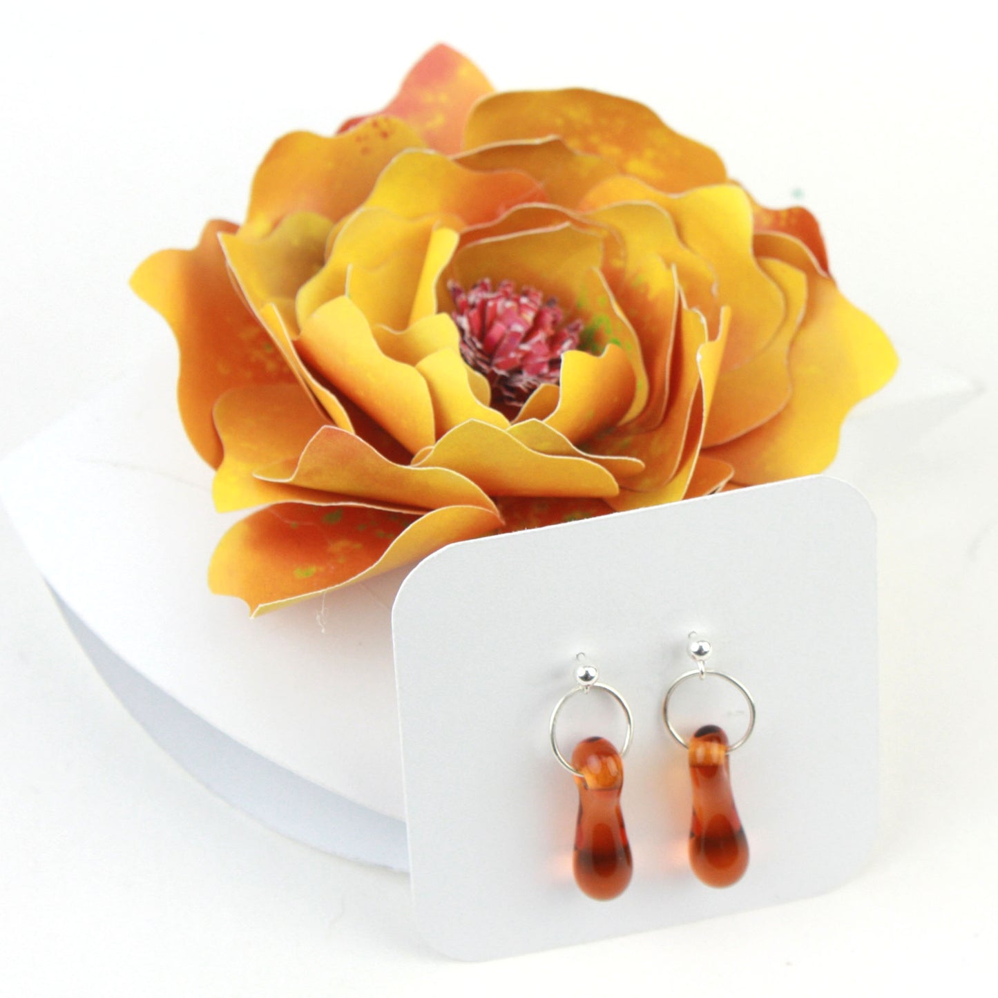 A pair of amber glass and sterling silver earrings displayed in a white gift box, adorned with a handcrafted golden-yellow paper flower.