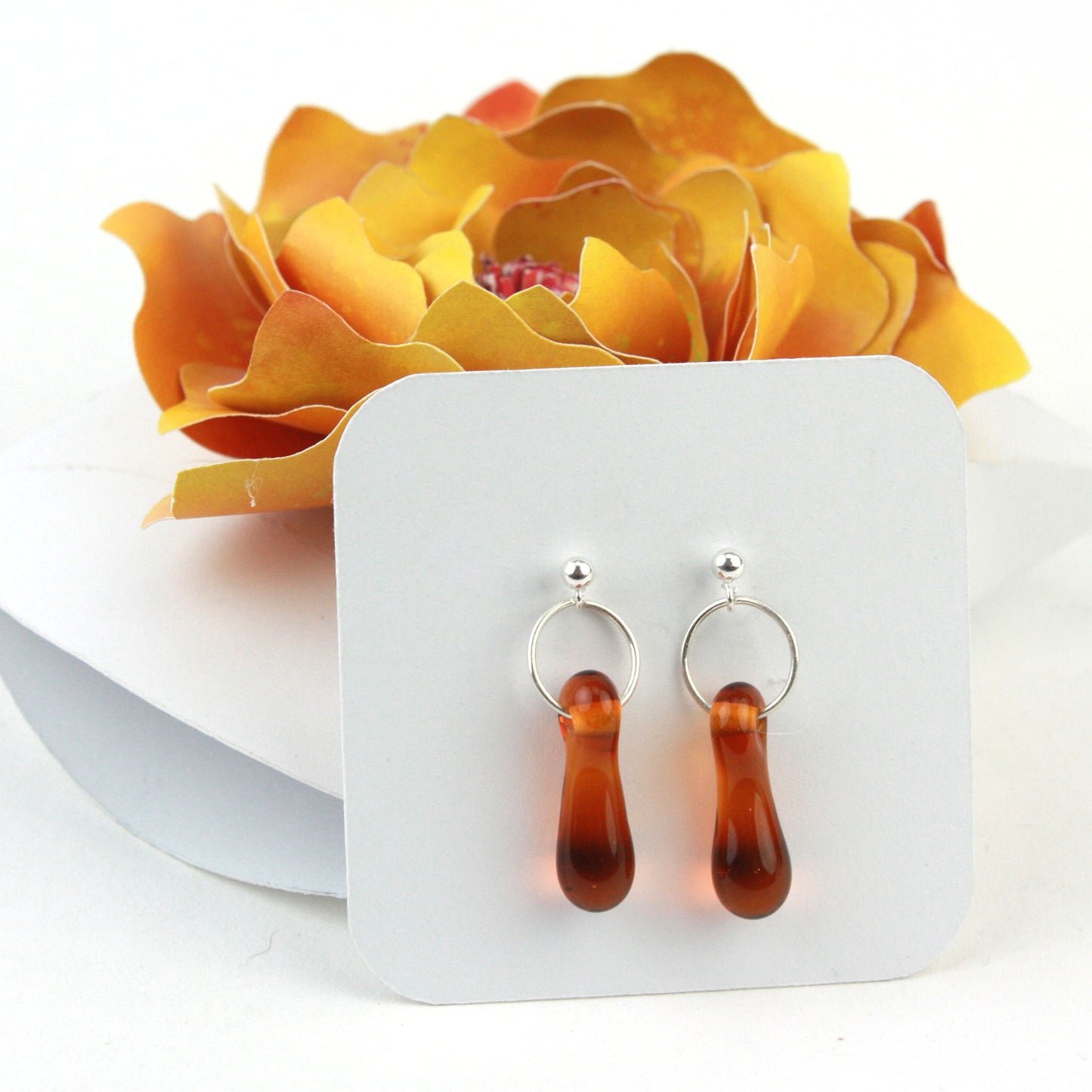 A pair of amber glass and sterling silver earrings displayed in a white gift box, adorned with a handcrafted golden-yellow paper flower.