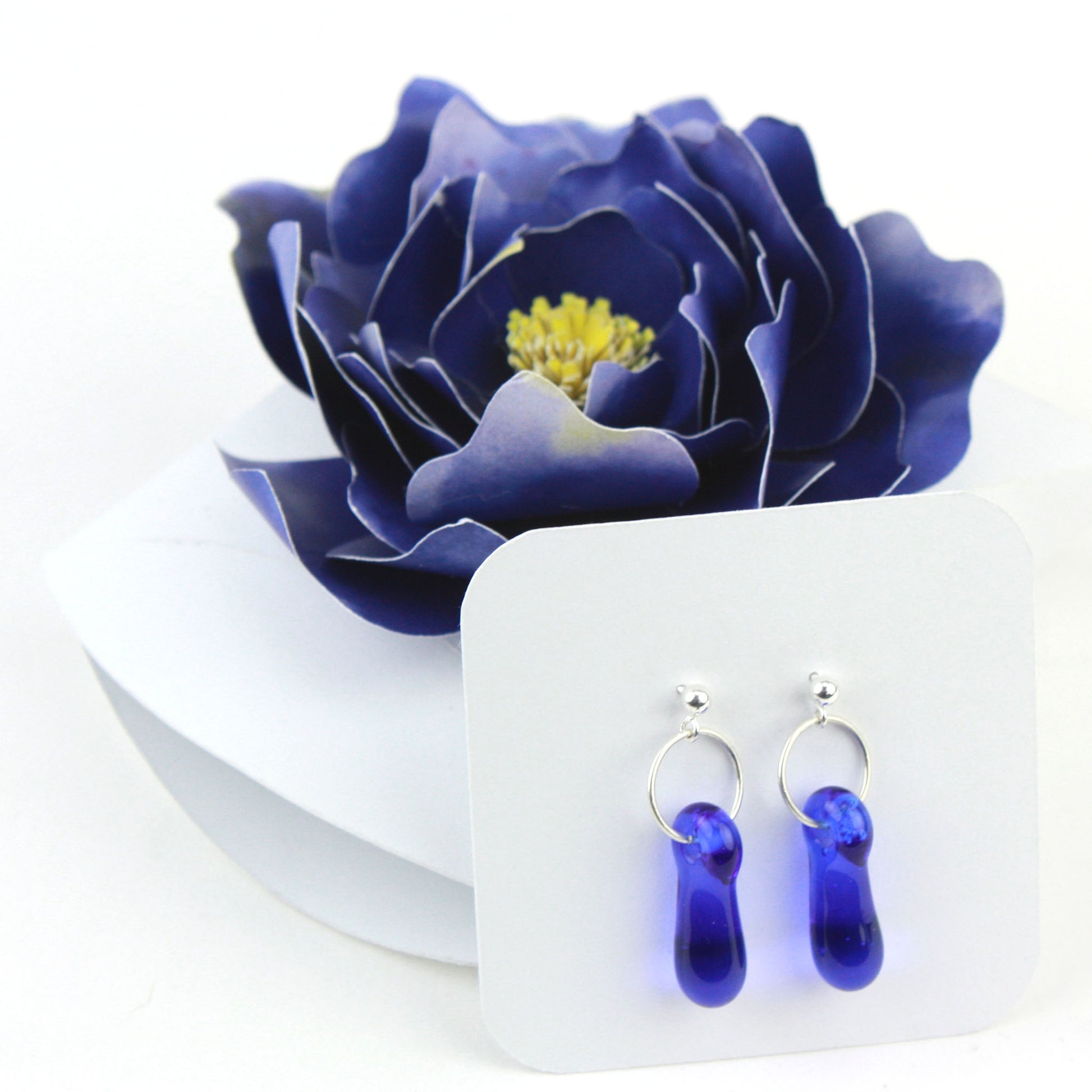 A pair of cobalt blue glass and sterling silver earrings displayed in a white gift box, decorated with a handcrafted deep blue paper flower.