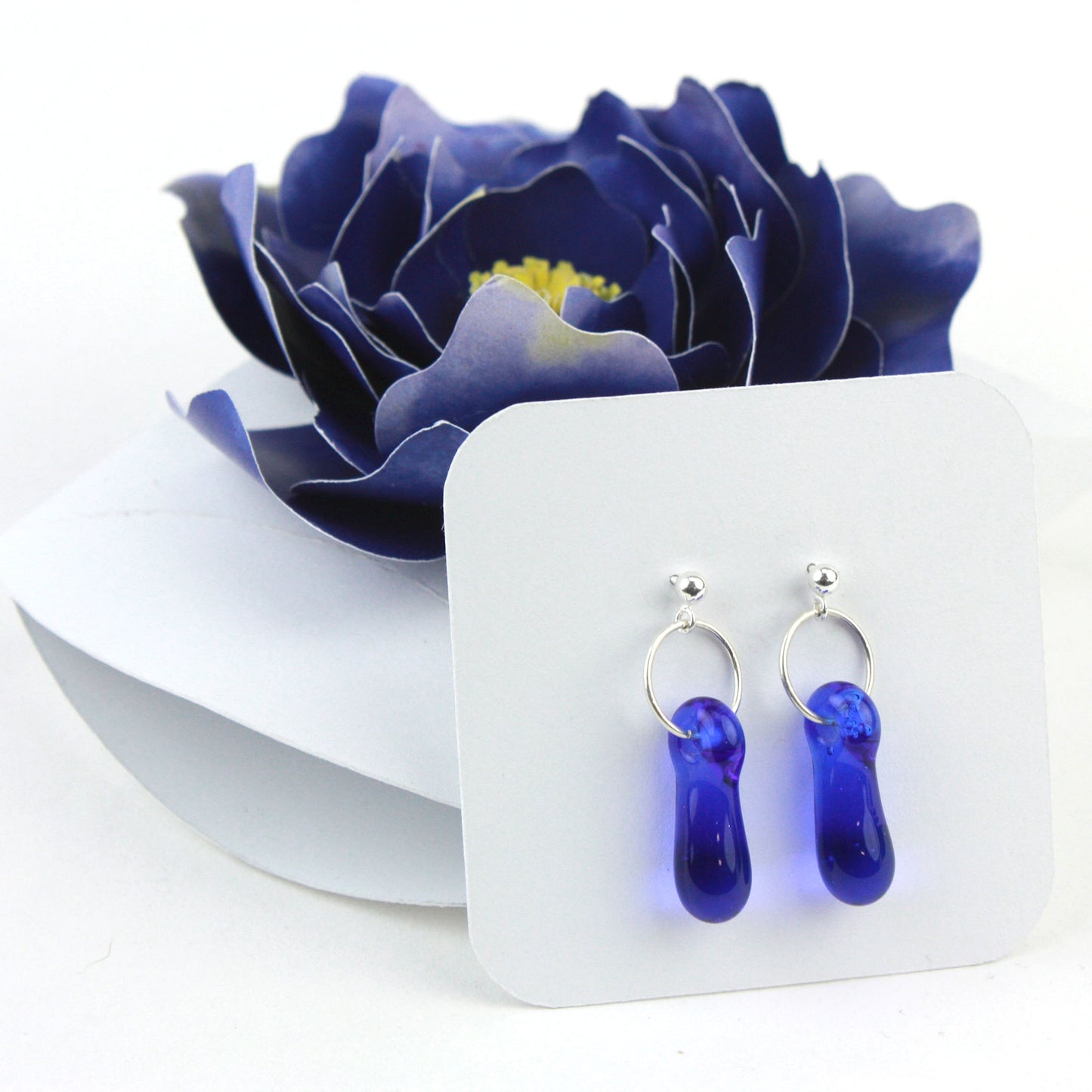 A pair of cobalt blue glass and sterling silver earrings displayed in a white gift box, decorated with a handcrafted deep blue paper flower.