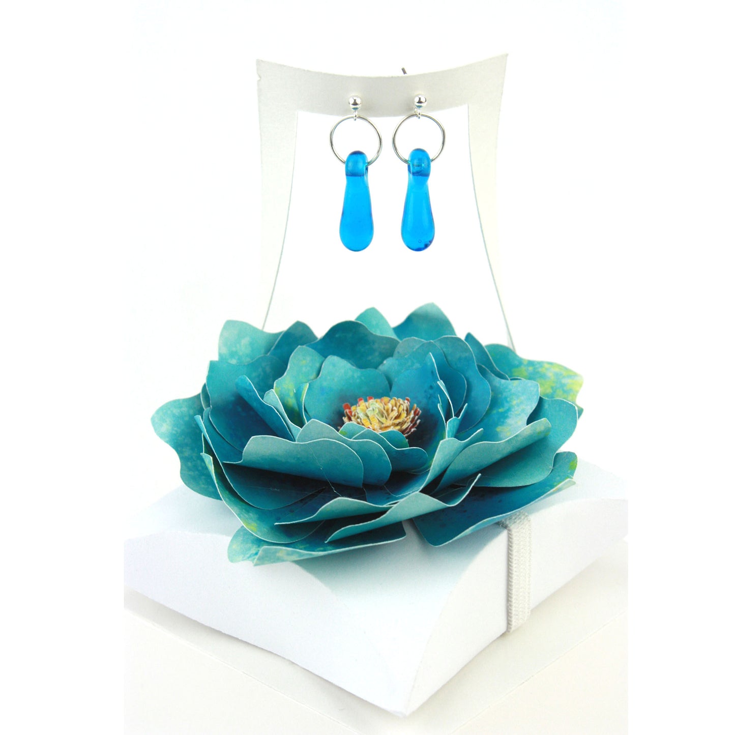 A pair of aqua blue glass and sterling silver earrings displayed in a white gift box, adorned with a handcrafted teal paper flower.