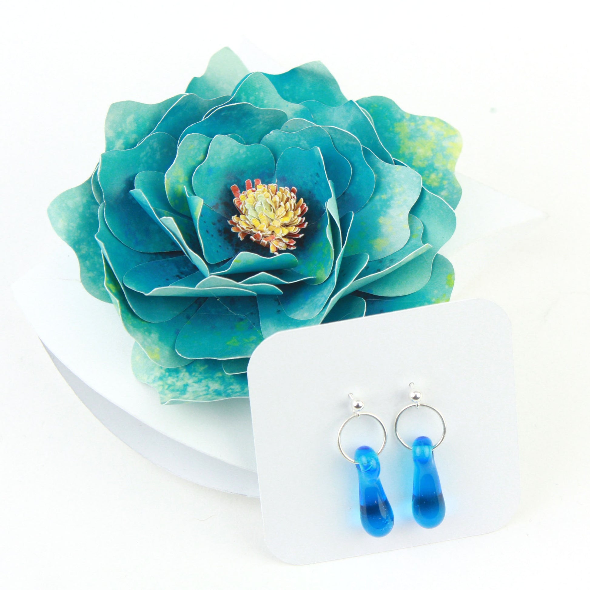 A pair of aqua blue glass and sterling silver earrings displayed in a white gift box, adorned with a handcrafted teal paper flower.