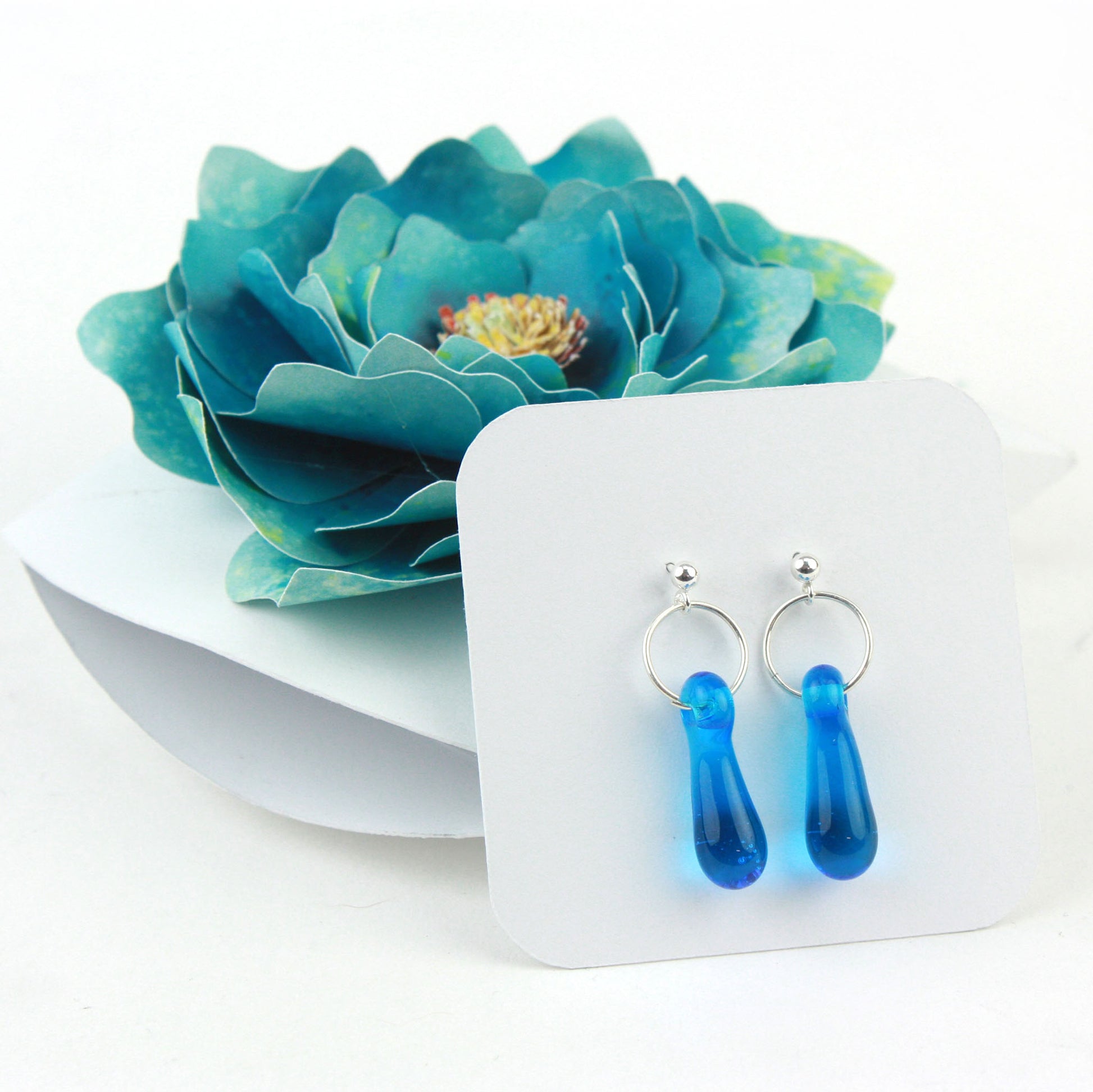 A pair of aqua blue glass and sterling silver earrings displayed in a white gift box, adorned with a handcrafted teal paper flower.