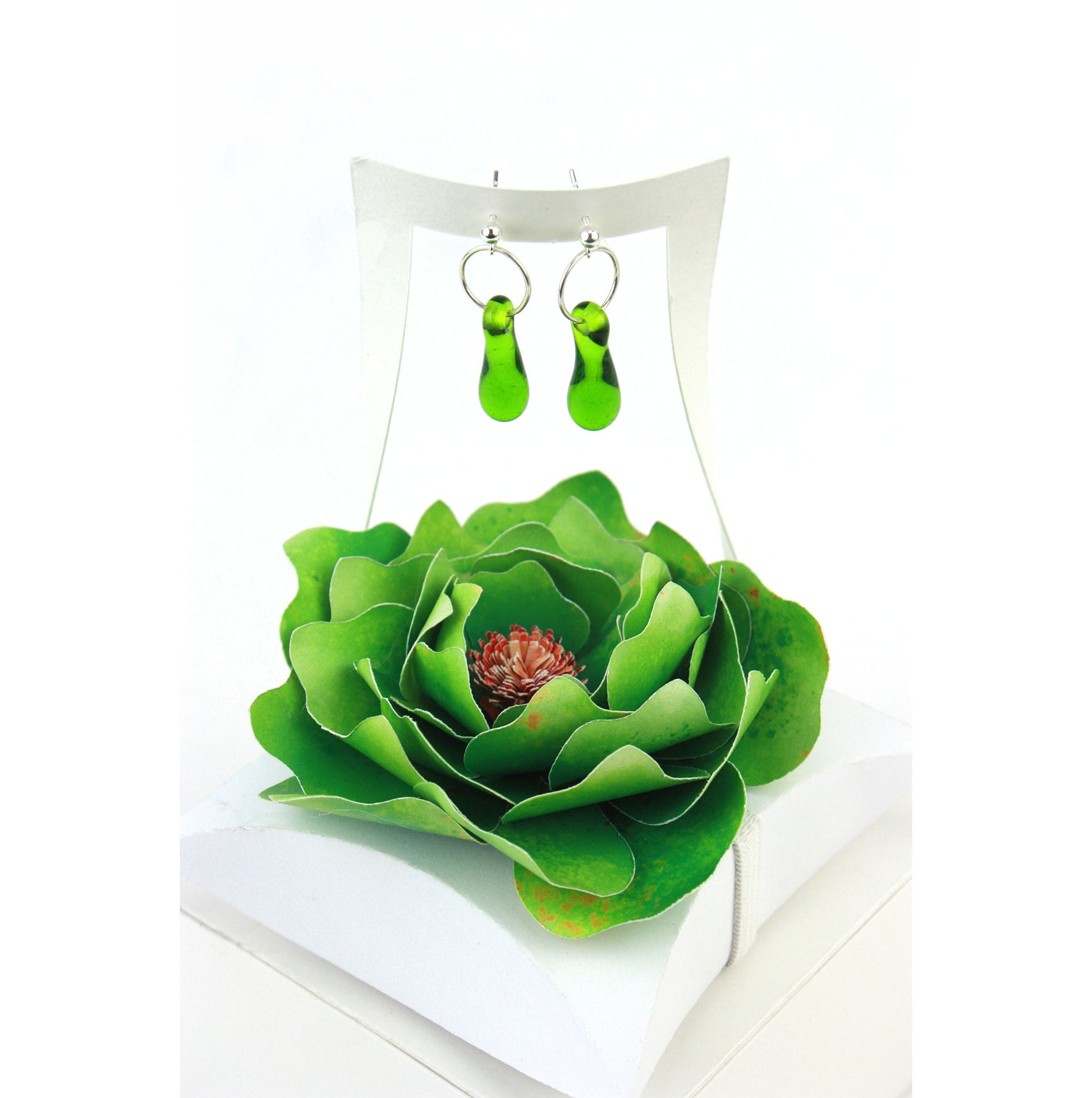 A pair of green glass and sterling silver earrings displayed in a white gift box, adorned with a handcrafted green paper flower.