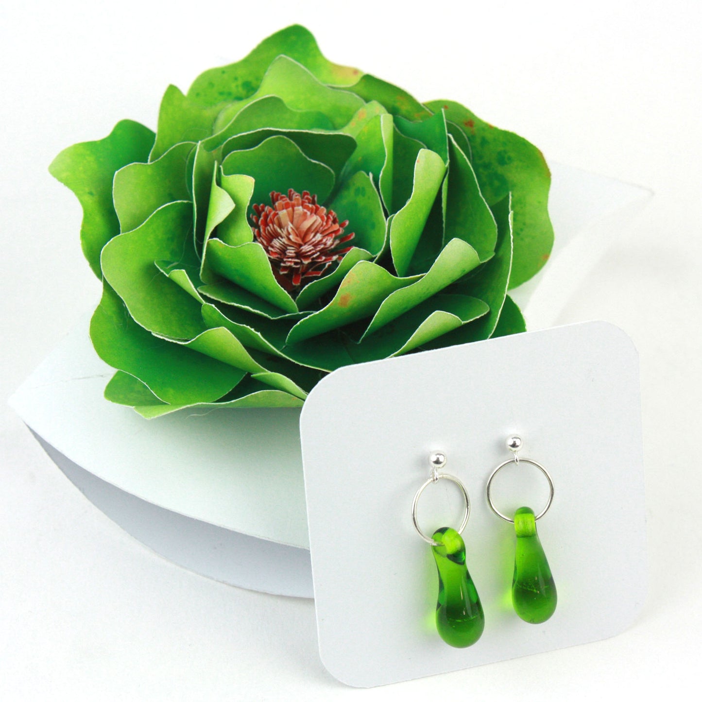 A pair of green glass and sterling silver earrings displayed in a white gift box, adorned with a handcrafted green paper flower.