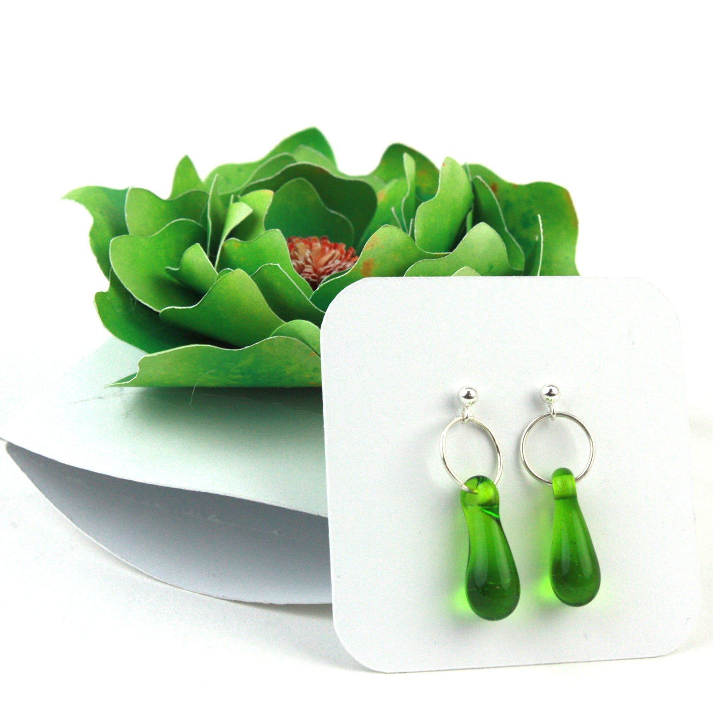 A pair of green glass and sterling silver earrings displayed in a white gift box, adorned with a handcrafted green paper flower.