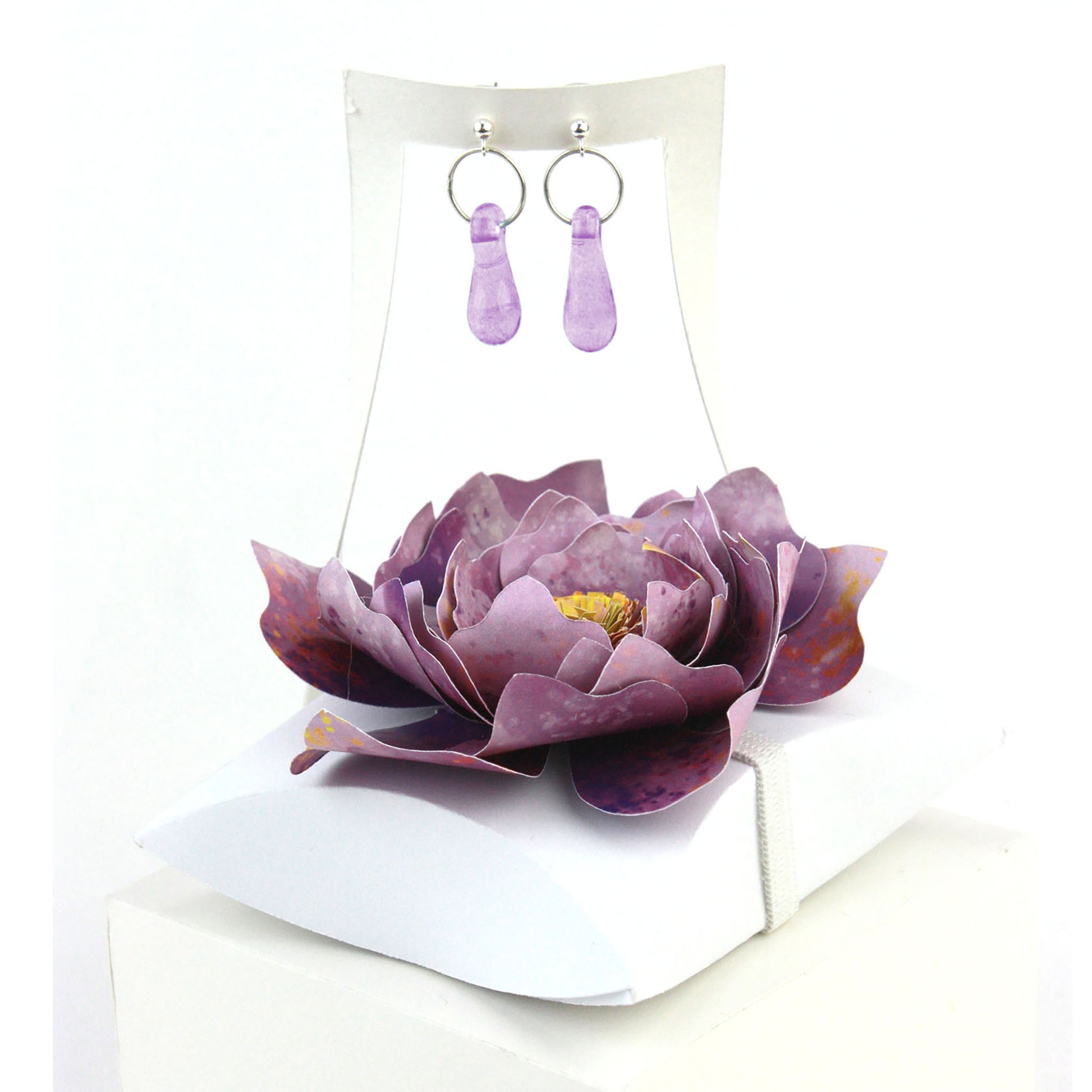 A pair of lavender purple glass and sterling silver earrings displayed in a white gift box, adorned with a handcrafted purple paper flower.