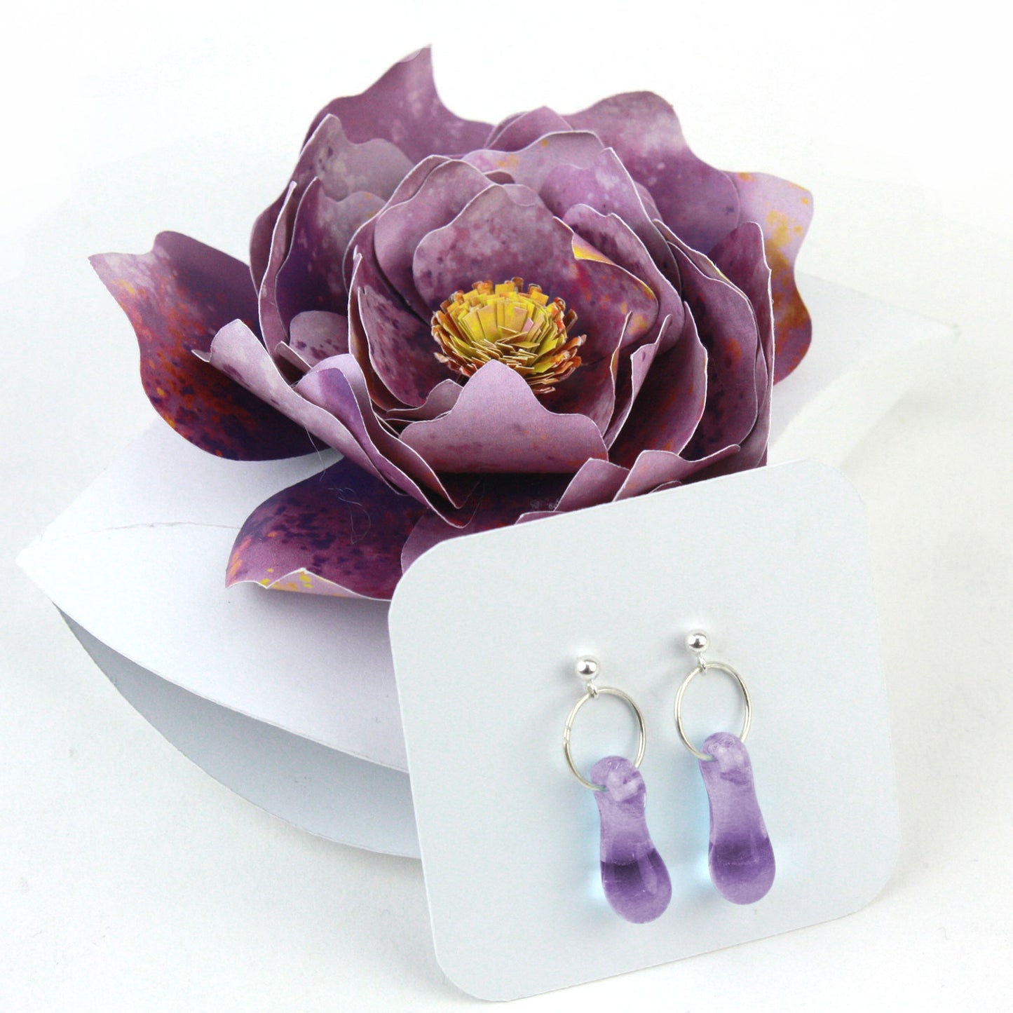 A pair of lavender purple glass and sterling silver earrings displayed in a white gift box, adorned with a handcrafted purple paper flower.