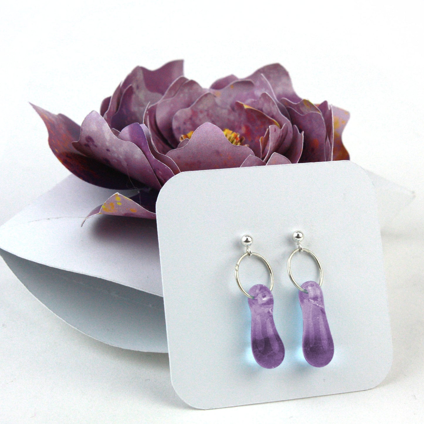 A pair of lavender purple glass and sterling silver earrings displayed in a white gift box, adorned with a handcrafted purple paper flower.