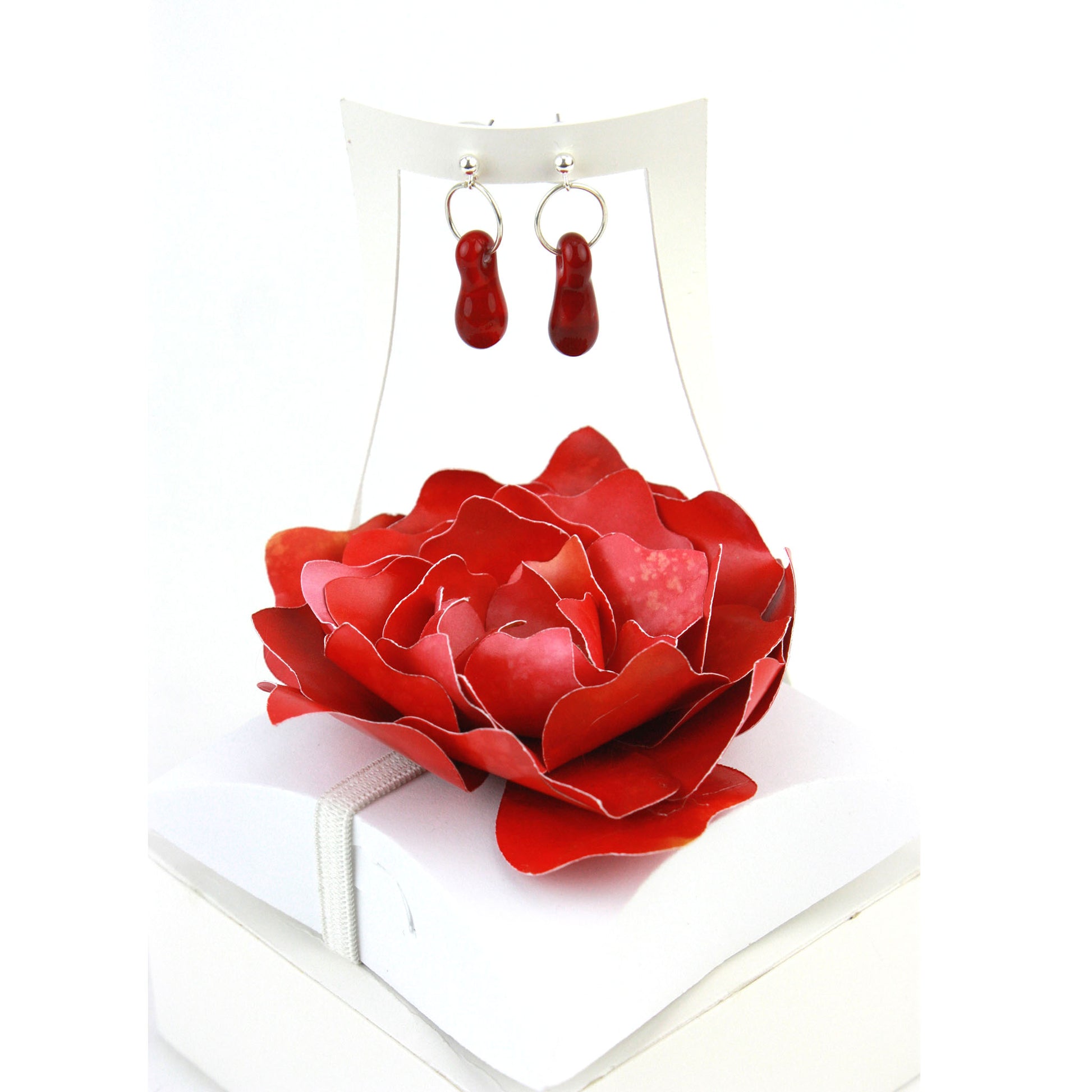 A pair of deep red glass and sterling silver earrings displayed in a white gift box, adorned with a handcrafted red paper flower.