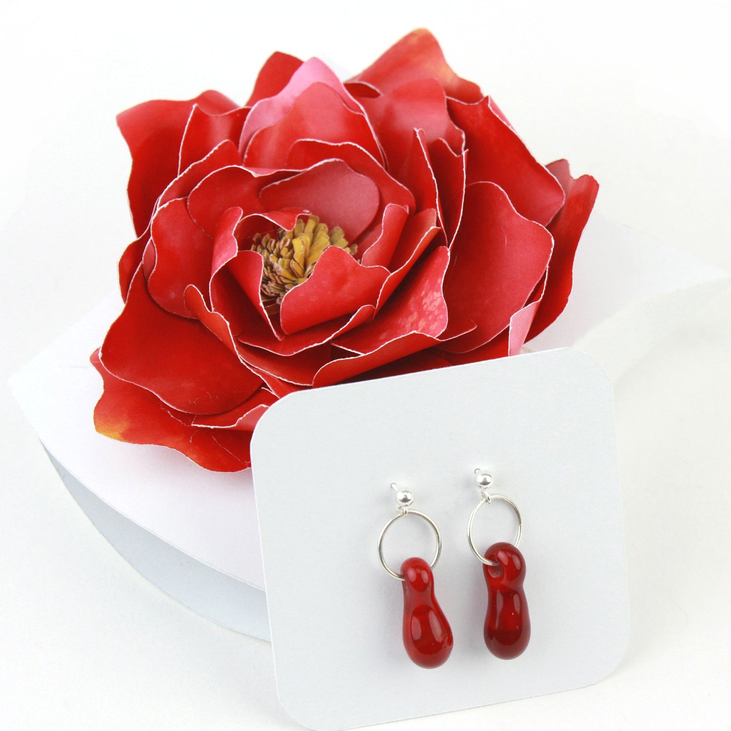 A pair of deep red glass and sterling silver earrings displayed in a white gift box, adorned with a handcrafted red paper flower.