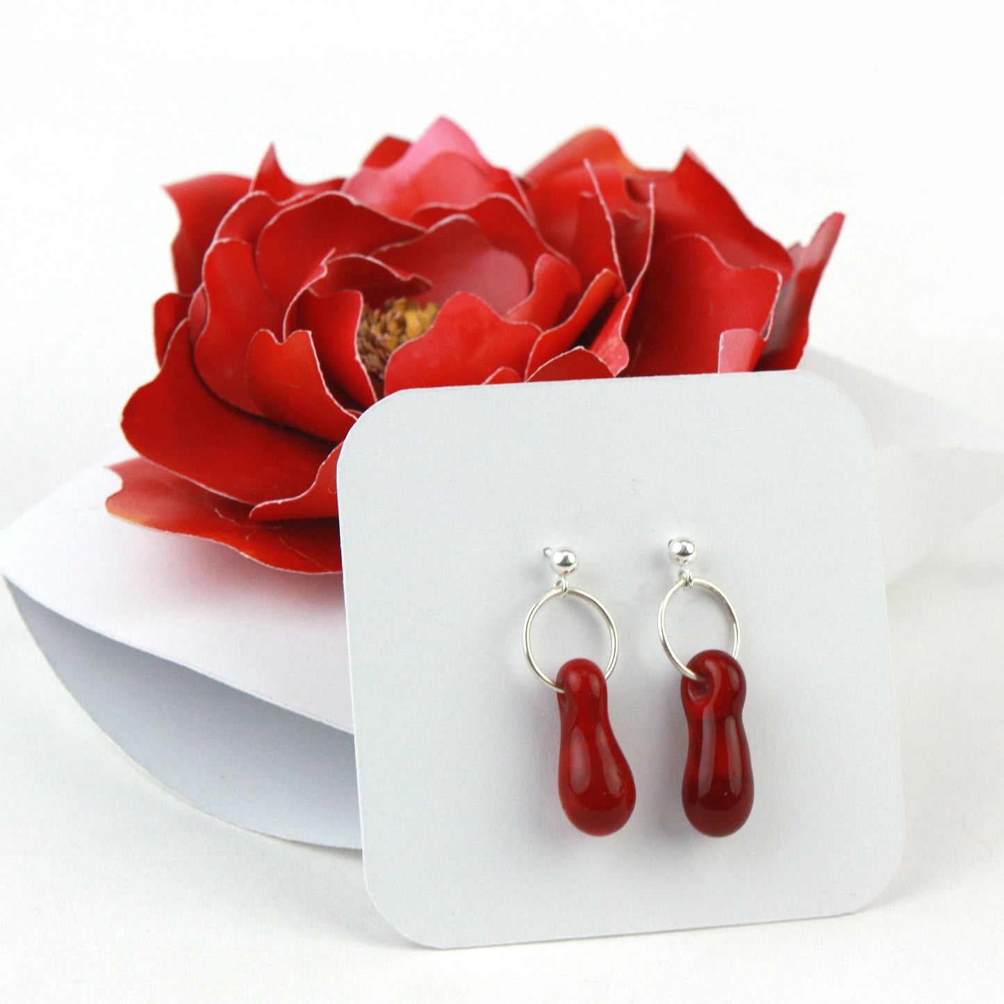 A pair of deep red glass and sterling silver earrings displayed in a white gift box, adorned with a handcrafted red paper flower.