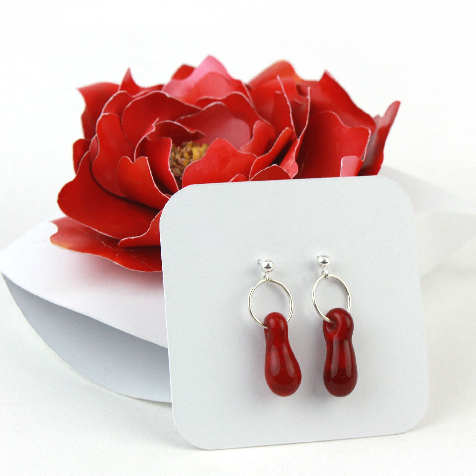 A pair of deep red glass and sterling silver earrings displayed in a white gift box, adorned with a handcrafted red paper flower.