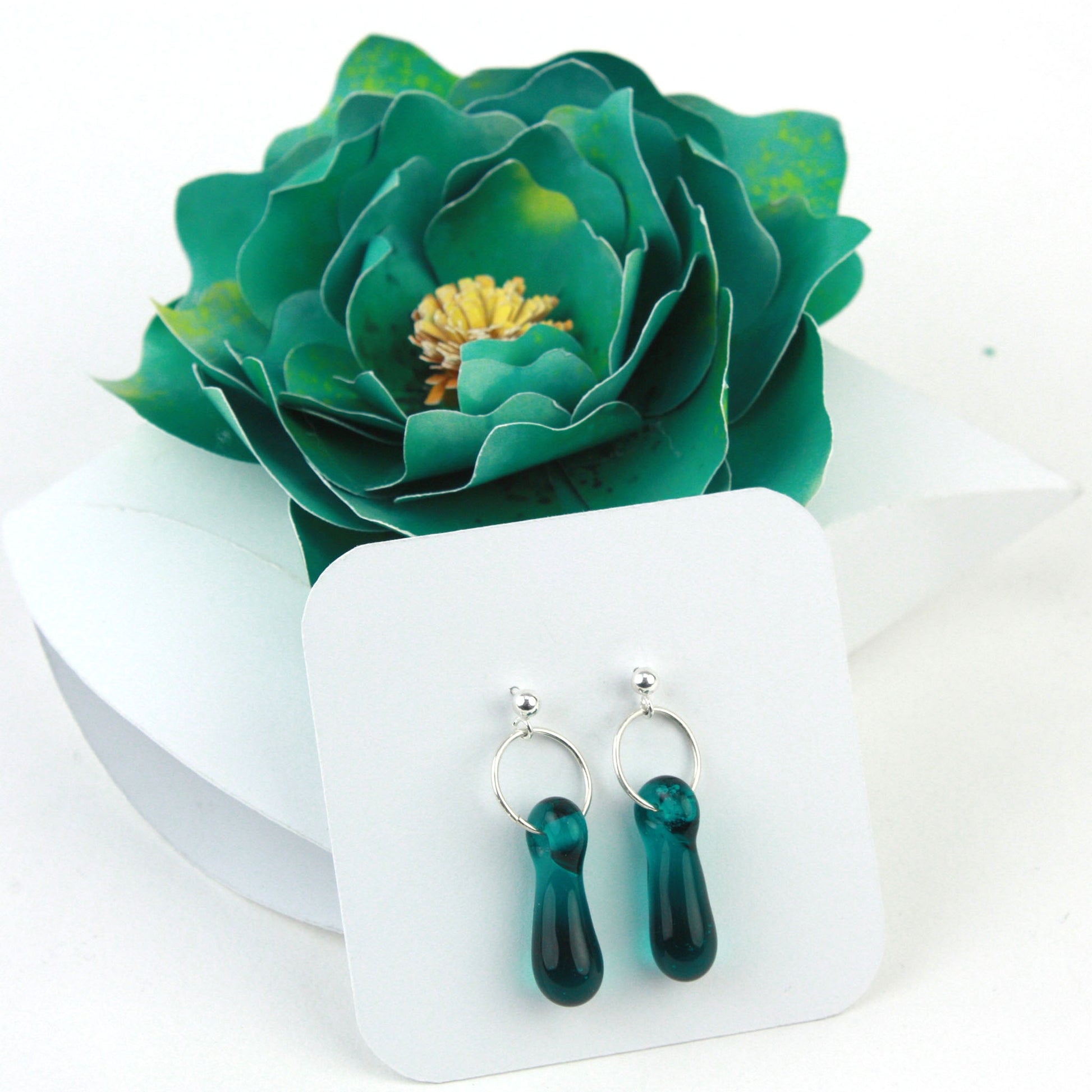 A pair of teal glass and sterling silver earrings displayed in a white gift box, adorned with a handcrafted teal paper flower.