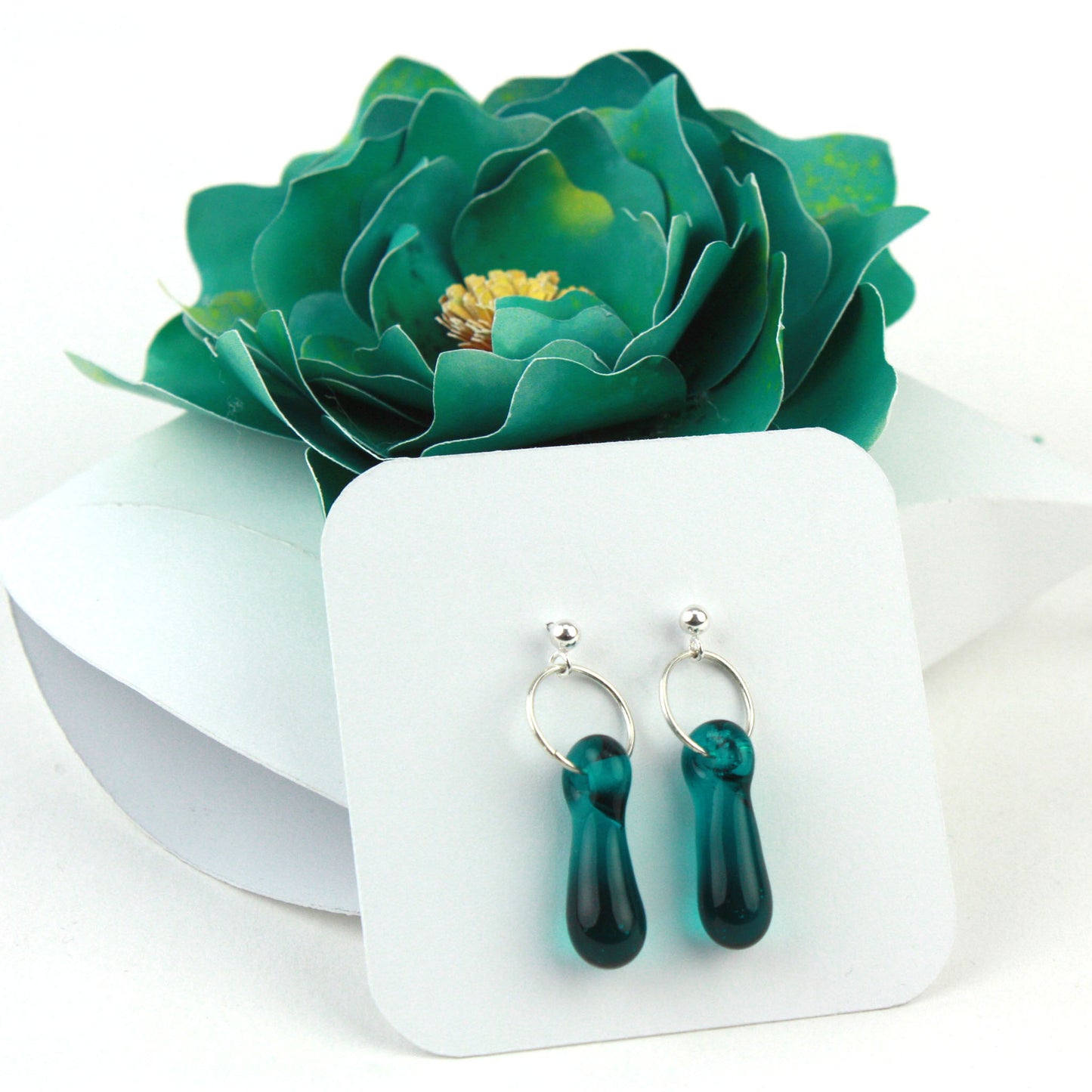 A pair of teal glass and sterling silver earrings displayed in a white gift box, adorned with a handcrafted teal paper flower.