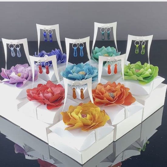 A collection of 12 handcrafted glass and sterling silver earrings, each in a white gift box with a colorful paper flower topper, designed for bulk retail restocking.